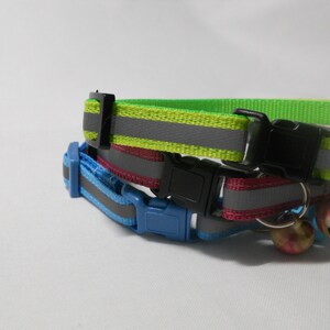 K&ouml;nnte beinhalten: Zwei Katzenhalsb&auml;nder mit reflektierenden Streifen. Eines ist neongr&uuml;n, das andere blau. Beide haben schwarze Kunststoffschnallen. Das gr&uuml;ne Halsband liegt auf einem rosa Halsband. An jedem Halsband ist eine kleine Glocke befestigt.