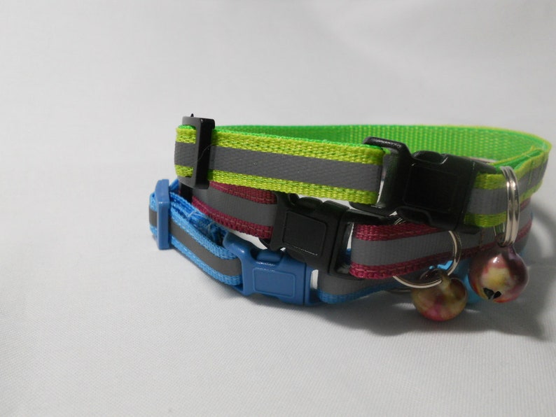 K&ouml;nnte beinhalten: Zwei Katzenhalsb&auml;nder mit reflektierenden Streifen. Eines ist neongr&uuml;n, das andere blau. Beide haben schwarze Kunststoffschnallen. Das gr&uuml;ne Halsband liegt auf einem rosa Halsband. An jedem Halsband ist eine kleine Glocke befestigt.