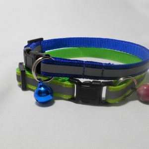 K&ouml;nnte beinhalten: Zwei Katzenhalsb&auml;nder. Eines ist blau und gr&uuml;n, das andere gr&uuml;n und gelb. Beide haben reflektierende Streifen, schwarze Kunststoffschnallen und eine kleine Glocke. Das blau-gr&uuml;ne Halsband hat eine blaue Glocke, das gr&uuml;n-gelbe eine rosarot-farbene Perle.