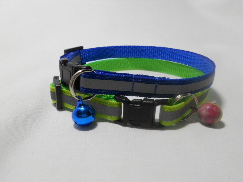 K&ouml;nnte beinhalten: Zwei Katzenhalsb&auml;nder. Eines ist blau und gr&uuml;n, das andere gr&uuml;n und gelb. Beide haben reflektierende Streifen, schwarze Kunststoffschnallen und eine kleine Glocke. Das blau-gr&uuml;ne Halsband hat eine blaue Glocke, das gr&uuml;n-gelbe eine rosarot-farbene Perle.