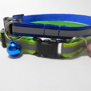 K&ouml;nnte beinhalten: Reflektierendes Katzenhalsband in Blau und Gr&uuml;n. Es hat eine schwarze Schnalle und einen silbernen Metallring mit einer blauen Glocke. Ein weiterer silberner Ring h&auml;lt eine rosa Glocke. Das Halsband hat einen reflektierenden Streifen.