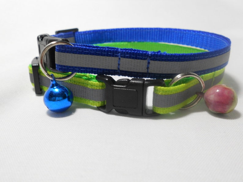 K&ouml;nnte beinhalten: Reflektierendes Katzenhalsband in Blau und Gr&uuml;n. Es hat eine schwarze Schnalle und einen silbernen Metallring mit einer blauen Glocke. Ein weiterer silberner Ring h&auml;lt eine rosa Glocke. Das Halsband hat einen reflektierenden Streifen.