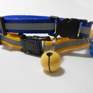 K&ouml;nnte beinhalten: Ein blau-gelbes Katzenhalsband mit reflektierendem Streifen und schwarzer Kunststoffschnalle. Das Halsband hat eine kleine gelbe Glocke und eine blaue Glocke. F&uuml;r Sicherheit und Sichtbarkeit konzipiert.