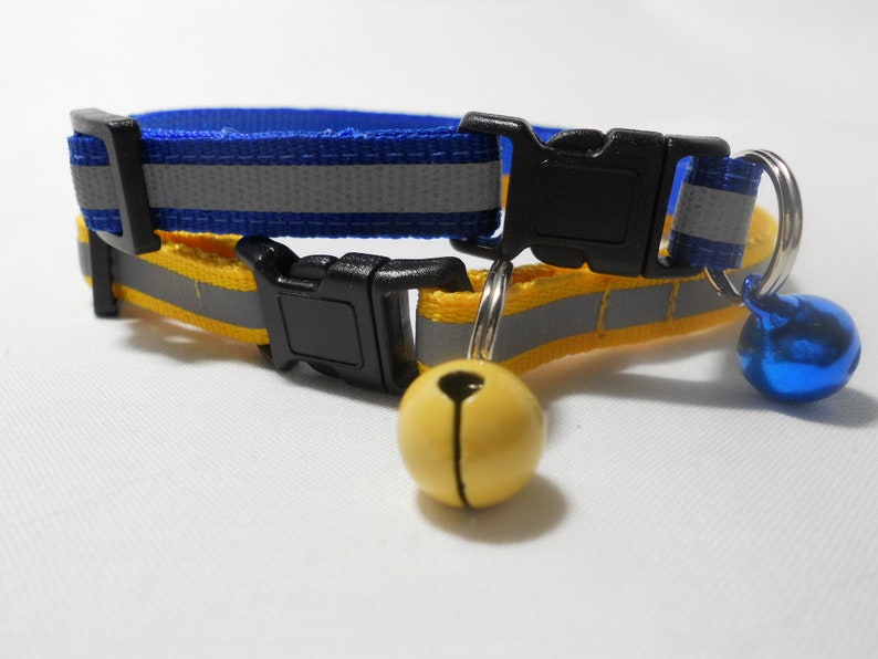 K&ouml;nnte beinhalten: Ein blau-gelbes Katzenhalsband mit reflektierendem Streifen und schwarzer Kunststoffschnalle. Das Halsband hat eine kleine gelbe Glocke und eine blaue Glocke. F&uuml;r Sicherheit und Sichtbarkeit konzipiert.