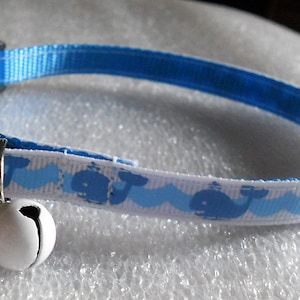 May include: A white and blue cat collar with a blue whale pattern and a white bell. The collar has a black plastic buckle.