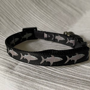 May include: Black pet collar with a repeating pattern of silver sharks. The collar has a black plastic buckle and a metal D-ring for leash attachment. The collar is made of a durable nylon material.