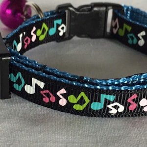 May include: A black pet collar featuring a repeating pattern of musical notes in shades of pink, green, and teal. The collar is constructed with a blue inner lining, a black plastic side-release buckle, and a small, round, metallic purple bell.