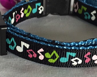 Könnte beinhalten: Ein schwarzes Haustierhalsband mit einem sich wiederholenden Muster aus Musiknoten in Rosa, Grün und Türkis. Das Halsband verfügt über ein blaues Innenfutter, eine schwarze Kunststoff-Steckschnalle und eine kleine, runde, metallisch violette Glocke.