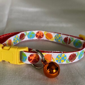 May include: A cat collar with a white band featuring colorful planet illustrations. The collar has a red trim, a yellow plastic buckle, and a small orange bell. The planets include Earth, Jupiter, and Saturn.