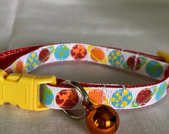 Könnte beinhalten: Ein Katzenhalsband mit einem weißen Band mit bunten Planetenillustrationen. Das Halsband hat eine rote Umrandung, eine gelbe Kunststoffschnalle und eine kleine orangefarbene Glocke. Zu den Planeten gehören Erde, Jupiter und Saturn.