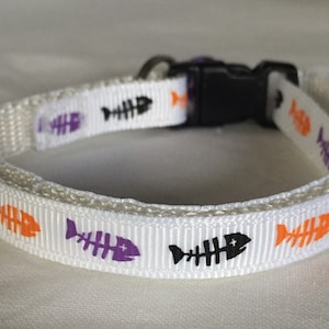 Puede incluir: Collar de gato blanco con un patrón repetido de espinas de pescado naranja y morado. El collar tiene una hebilla de plástico negra.