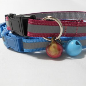 K&ouml;nnte beinhalten: Zwei Katzenhalsb&auml;nder, eines grau und bordeauxrot, das andere blau. Beide haben reflektierende Streifen und schwarze oder blaue Kunststoffschnallen. Jedes Halsband hat eine kleine Glocke, eine gold-rot, die andere blau.