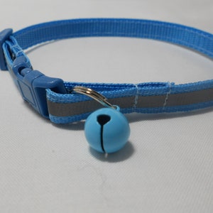 K&ouml;nnte beinhalten: Ein hellblaues Katzenhalsband mit einem reflektierenden Streifen und einer passenden blauen Kunststoffschnalle. Eine kleine, hellblaue Glocke h&auml;ngt an einem silberfarbenen Ring. Das Halsband ist f&uuml;r die Sicherheit und Sichtbarkeit von Haustieren konzipiert.