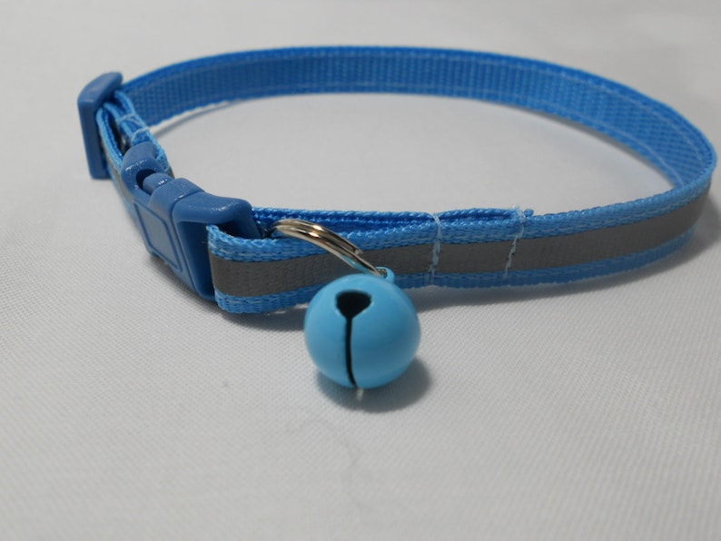 K&ouml;nnte beinhalten: Ein hellblaues Katzenhalsband mit einem reflektierenden Streifen und einer passenden blauen Kunststoffschnalle. Eine kleine, hellblaue Glocke h&auml;ngt an einem silberfarbenen Ring. Das Halsband ist f&uuml;r die Sicherheit und Sichtbarkeit von Haustieren konzipiert.