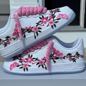 May include: A pair of white sneakers featuring thick pink rope laces and embroidered floral designs with pink blossoms, green stems, and pink butterflies. The shoes have a white base, a small pink swoosh logo, and the text AIR embossed on the midsole.