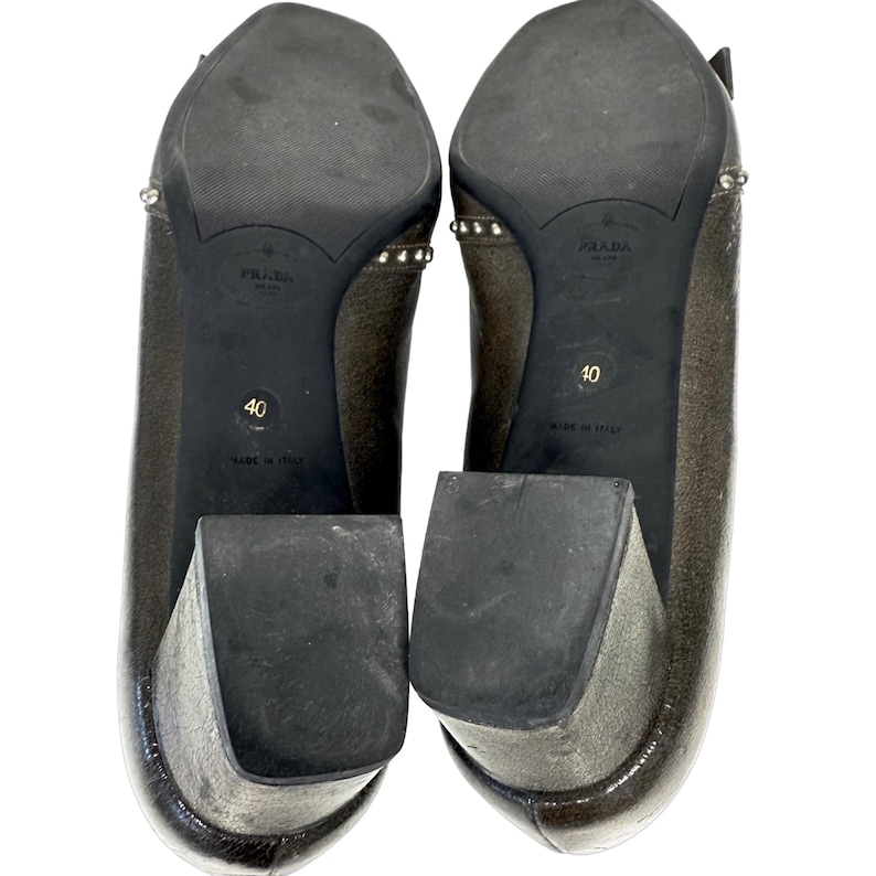 May include: Pair of metallic bronze-colored Prada heels, size 40, with block heels and studded accents. The soles are black with the Prada logo and "Made in Italy" text.