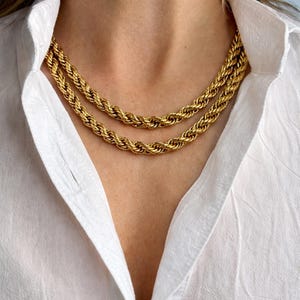 May include: Two gold-colored rope chain necklaces layered on a person's neck. The necklaces are thick and have a twisted design. The person is wearing a white button-down shirt.