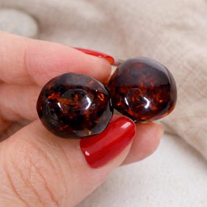 Puede incluir: Dos pendientes redondos de color ámbar oscuro con un acabado brillante. Los pendientes tienen un aspecto veteado, con tonos más claros y oscuros de marrón y naranja. Los pendientes se sostienen entre los dedos con esmalte de uñas rojo.