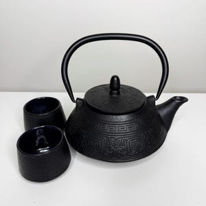 Pode incluir: Uma chaleira de ferro fundido preta com uma alça curva e duas xícaras combinando. A chaleira tem uma superfície texturizada com um padrão decorativo. As xícaras são pequenas e azul escuro.