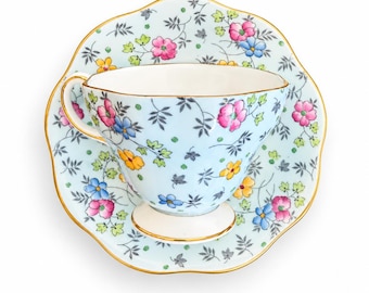 May include: A light blue teacup and matching saucer set featuring a delicate floral pattern with pink, yellow, and blue flowers, green leaves, and dark gray foliage. Both pieces have a scalloped edge and are trimmed with a thin gold rim.