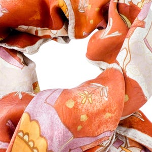 May include: A scarf featuring a vibrant orange and cream color palette with a repeating pattern of stylized flowers, leaves, and geometric shapes. The fabric has a smooth, slightly lustrous texture with visible woven lines throughout the material.