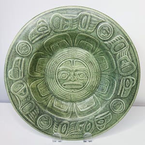 May include: A green ceramic plate with a detailed, embossed design. The central image is a stylized sun face, surrounded by a ring of geometric and animal-like figures. The plate has a speckled texture and is displayed on a clear stand.