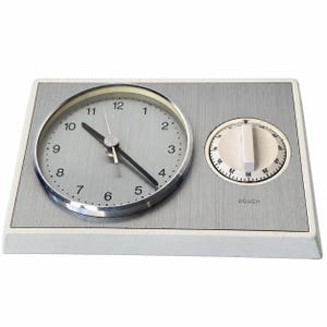 May include: A rectangular Bosch kitchen timer and clock combination with a white plastic base and a brushed silver metal face. The left side features an analog clock with black hands and numerals, while the right side has a circular timer dial.
