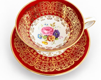 May include: A porcelain teacup and saucer set featuring a deep red background with intricate gold filigree patterns. The center of the cup displays a colorful floral bouquet with pink, yellow, and blue flowers on a white base. The handle is white.