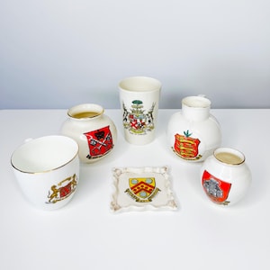 May include: A collection of white ceramic items, including cups, vases, and a small dish, each adorned with colorful heraldic crests. The items vary in shape and size, with gold trim on some. The crests feature coats of arms.