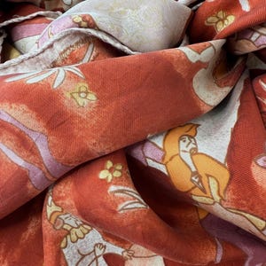 May include: A rust-orange fabric scarf featuring a repeating pattern of stylized figures in yellow and white robes, accented with small yellow flowers and light pink geometric shapes. The material has a visible ribbed texture and a finished hem.