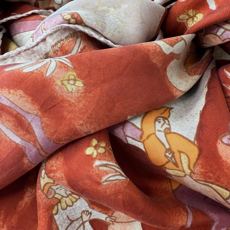 May include: A rust-orange fabric scarf featuring a repeating pattern of stylized figures in yellow and white robes, accented with small yellow flowers and light pink geometric shapes. The material has a visible ribbed texture and a finished hem.