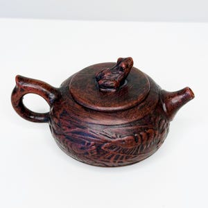 Puede incluir: Pequeña tetera de cerámica marrón con tapa y asa. La tetera tiene una superficie texturizada con detalles tallados. La tapa presenta un asa en forma de rana. Diseñada para preparar té.
