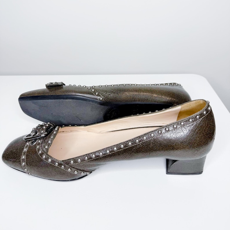 May include: A pair of brown leather pumps with silver-tone stud accents. The shoes have a square toe, a small block heel, and a decorative buckle. One shoe is positioned on top of the other.
