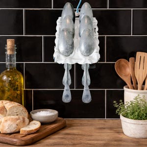 May include: A kitchen scene featuring a white spoon rack holding six silver spoons. The spoons have ornate handles. Other items include a bottle of olive oil, bread, salt, and wooden utensils. The background is a black tiled wall.