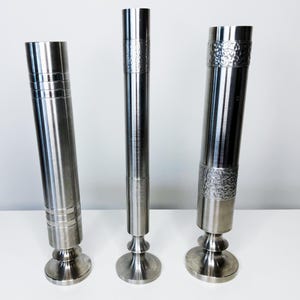 Puede incluir: Tres candelabros de metal plateado con diferentes texturas y diseños. Los candelabros tienen forma cilíndrica con bandas decorativas y secciones texturizadas. Cada uno tiene una base redonda y una parte superior acampanada, adecuada para velas.