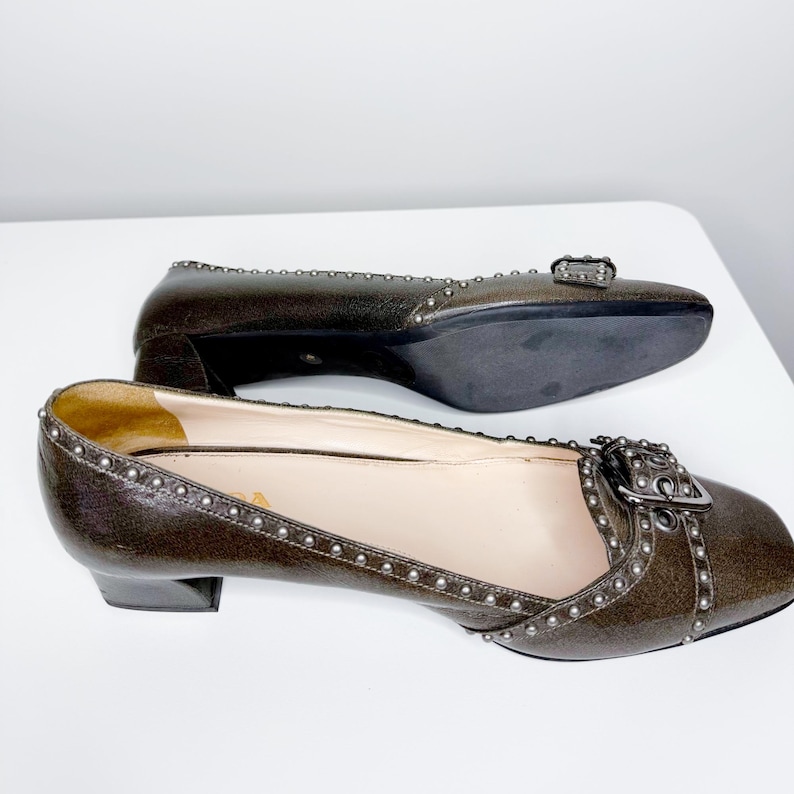 May include: A pair of brown leather flats with a square toe and small heel. The shoes feature a decorative buckle and are embellished with small silver studs along the edges.
