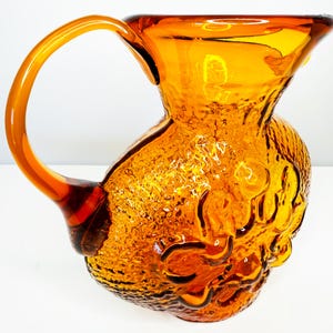 Puede incluir: Jarra de vidrio color ámbar con superficie texturizada y asa curva. La jarra tiene una abertura ancha y un diseño decorativo en el cuerpo. Una pieza decorativa de estilo vintage.