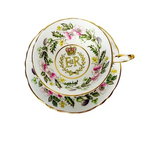 Op de afbeelding: Witte theekop en schotel set met een bloemenontwerp in roze, groen en geel. De theekop en schotel hebben een gouden rand. Het midden van de theekop heeft een kroon en de gouden letters "ER".