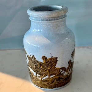 Puede incluir: Un pequeño tarro de cerámica azul claro con un esmalte craquelado. El tarro está decorado con una escena dorada de personas a caballo cazando.