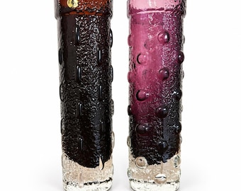 May include: Two cylindrical glass vases with a textured, pebbled surface. The left vase is dark brown, and the right vase is deep purple. Both feature a clear, bubble-textured base and a gold circular sticker on the upper rim of the brown vase.