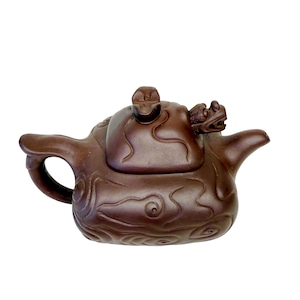 Op de afbeelding: Een bruine keramische theepot met een drakenontwerp op de tuit en het deksel. De theepot heeft een gebogen handvat en een decoratief wervelpatroon op de body. Het deksel heeft een kleine draakvormige knop.