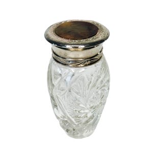 May include: A clear glass perfume bottle with a silver-coloured metal rim and lid. The bottle has a textured, starburst pattern. The lid is a dark brown colour. The bottle is empty and sits on a white background.