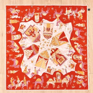 May include: A square silk scarf featuring a vibrant red border with a repeating pattern of figures riding camels. The central white area displays four ornate, tent-like structures in shades of orange, pink, and gold, arranged in a radial design.