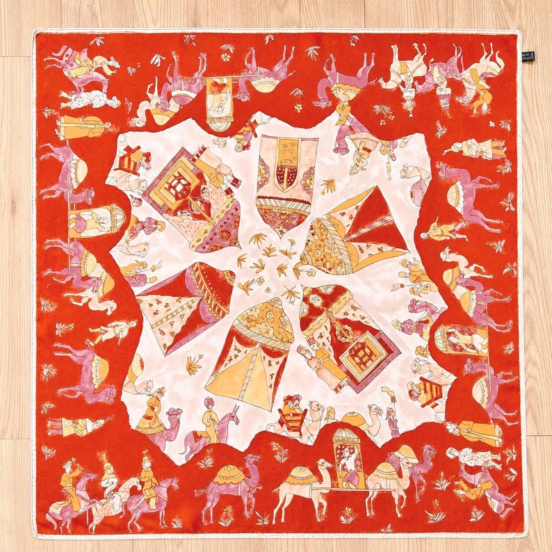 May include: A square silk scarf featuring a vibrant red border with a repeating pattern of figures riding camels. The central white area displays four ornate, tent-like structures in shades of orange, pink, and gold, arranged in a radial design.