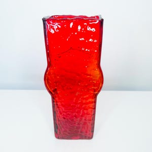 May include: A vibrant red glass vase with a textured surface. The vase has a unique shape, wider in the middle and narrower at the top and bottom. The rim has a wavy design. The vase is likely used for decorative purposes.