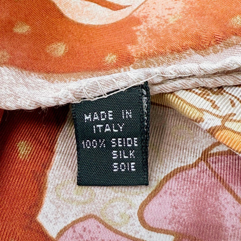 May include: A silk scarf featuring a vibrant orange, pink, and cream patterned fabric with a stitched hem. A black rectangular tag is attached, displaying the white text: MADE IN ITALY, 100% SEIDE, SILK, SOIE, indicating the origin and material.