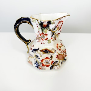 May include: A white ceramic pitcher with a floral design featuring red, blue, and gold accents. The handle is dark blue with gold trim. The pitcher has a unique shape and is likely vintage.