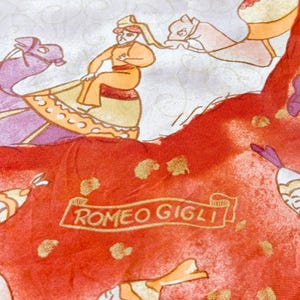 May include: A silk scarf featuring a vibrant red section with gold speckles and the text ROMEO GIGLI printed on a banner. The design includes illustrations of figures riding camels in shades of orange, purple, and cream against a light background.