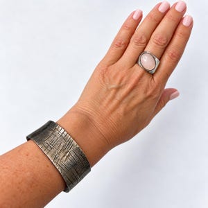 May include: A wide, dark metal cuff bracelet featuring a textured, cross-hatched surface pattern and a silver-toned ring set with a smooth, oval-shaped, pale pink gemstone worn on the finger.