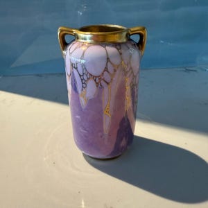 Puede incluir: Un pequeño jarrón de cerámica morado y blanco con detalles dorados y dos asas. El jarrón tiene un diseño único y abstracto con una superficie texturizada.